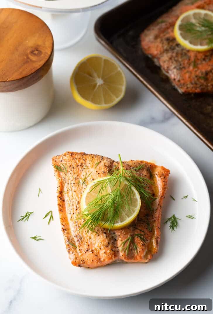 Perfect Oven-Baked Salmon 2 Top down view of a large filet of salmon topped with a lemon slice and sprigs of fresh dill. The fish is on a white plate.