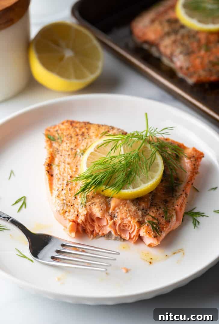 Perfect Oven-Baked Salmon 11 Large filet of salmon topped with a lemon slice and sprigs of fresh dill. The fish is on a white plate. A fork has removed a bite from the salmon.