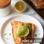 Top down view of a large filet of salmon topped with a lemon slice and sprigs of fresh dill. The fish is on a white plate.