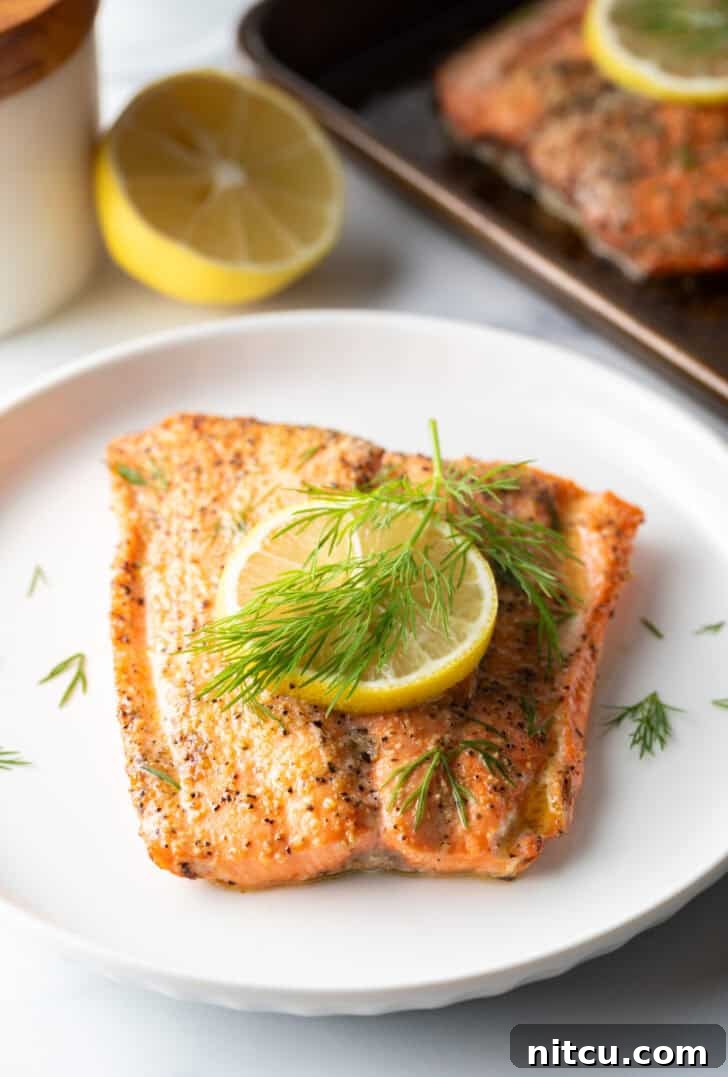 Perfect Oven-Baked Salmon 4 Large filet of salmon topped with a lemon slice and sprigs of fresh dill. The fish is on a white plate.