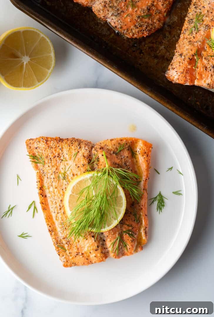 Perfect Oven-Baked Salmon 10 Top down view of a large filet of salmon topped with a lemon slice and sprigs of fresh dill. The fish is on a white plate.