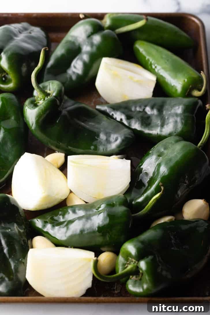 Whole poblano peppers, large chunks of sweet onion, and several cloves of garlic artfully arranged on a baking sheet, ready for roasting.