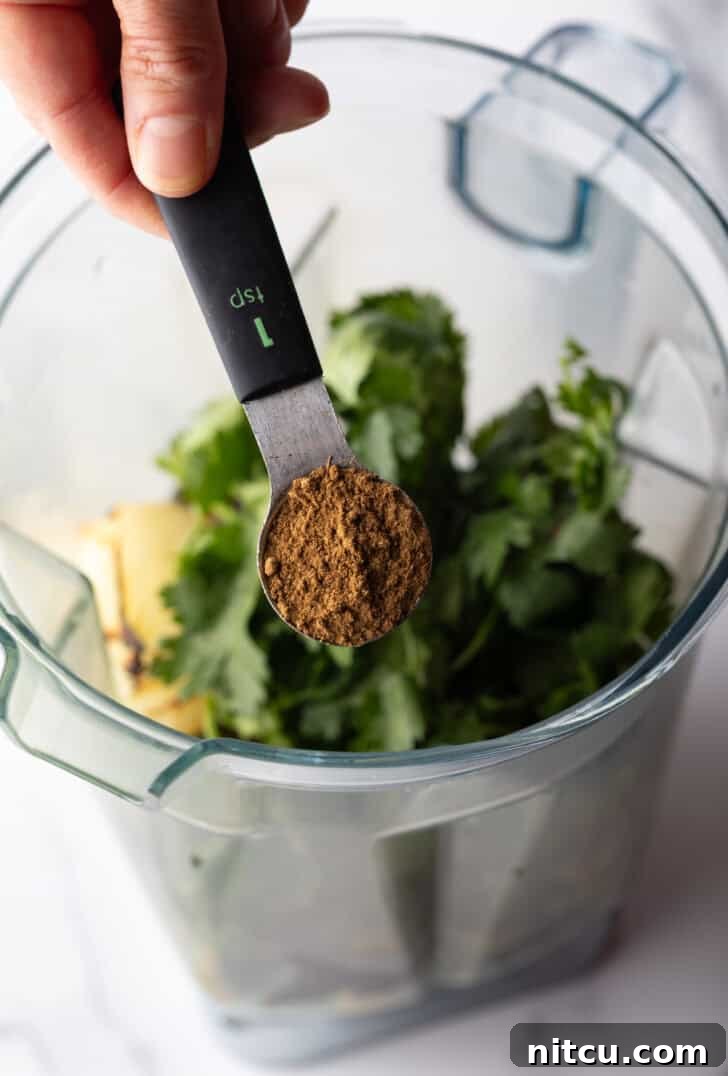 A hand holds a measuring spoon, pouring ground cumin into a clear blender pitcher filled with vibrant green herbs and roasted vegetables.