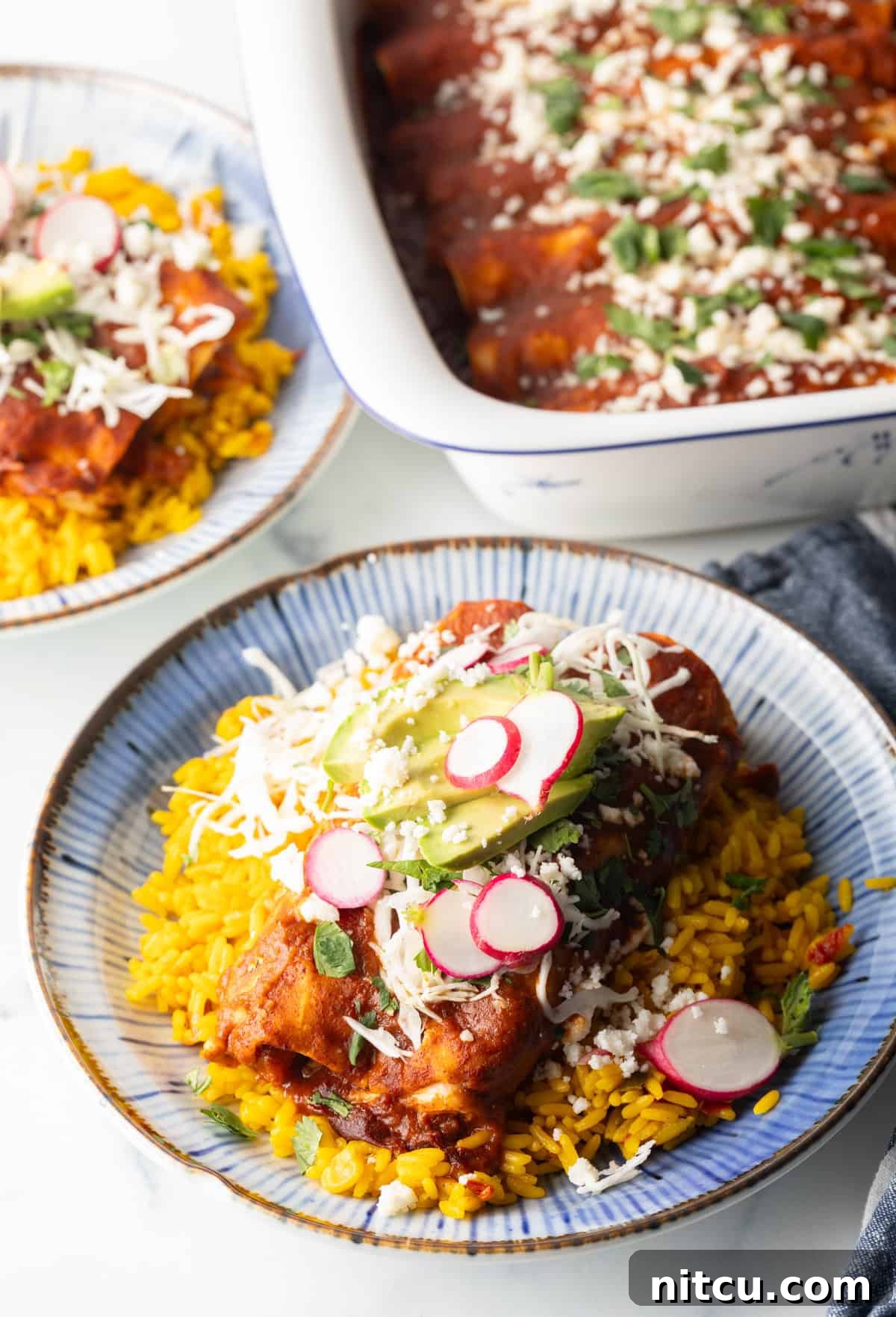 Authentic Red Enchilada Sauce 11 Large plateful of enchiladas with red sauce, topped with slices of radishes and fresh slaw, on a plate next to yellow rice. A large white baking dish of enchiladas is in the background.