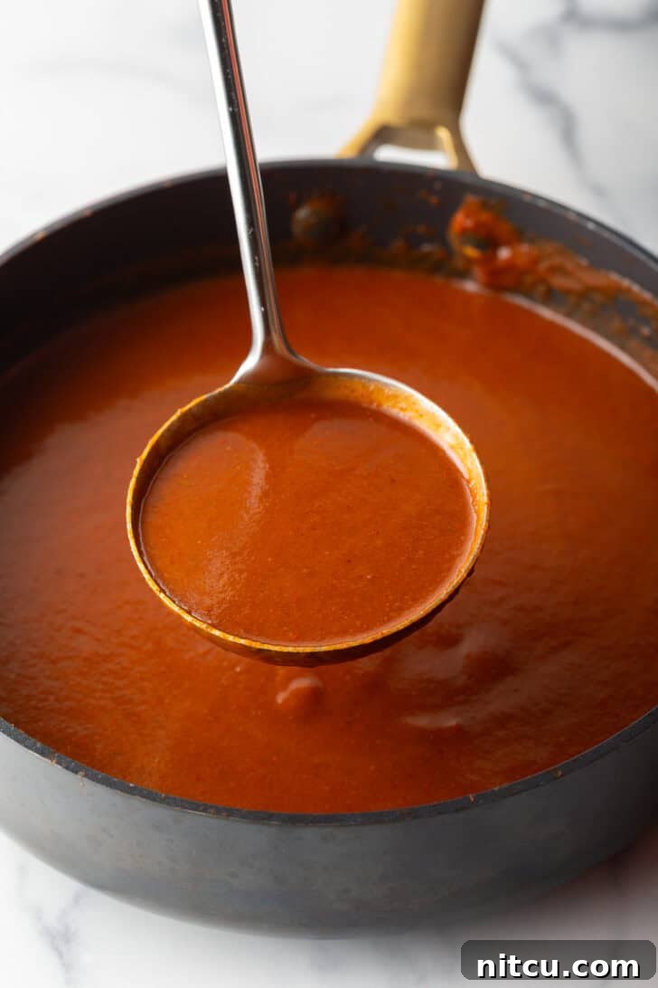 Authentic Red Enchilada Sauce 8 Metal ladleful of red sauce being held above a black pan of sauce.