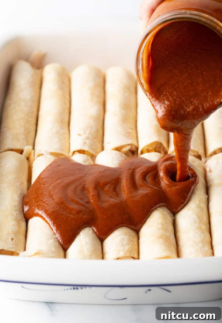 Grandma's Secret Enchiladas 8 Pouring red sauce from a glass jar over the top of a dozen rolled enchiladas in a white baking dish.
