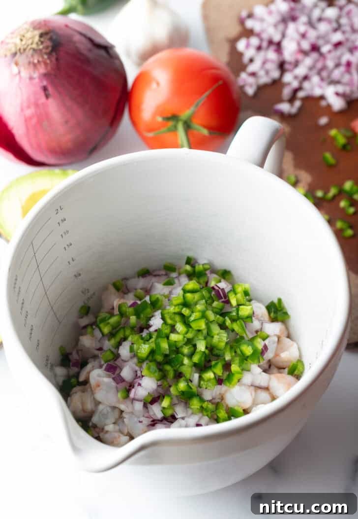 A white bowl featuring raw shrimp meticulously arranged, topped with a colorful scattering of finely chopped red and green peppers, showcasing the initial stage of ceviche preparation.