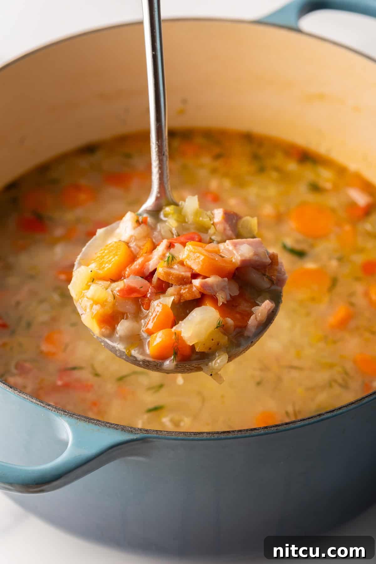 Comforting Ham and Cabbage Pot 6 Metal ladle holding a scoopful of cabbage ham soup to camera, held over a blue cooking pot of soup.