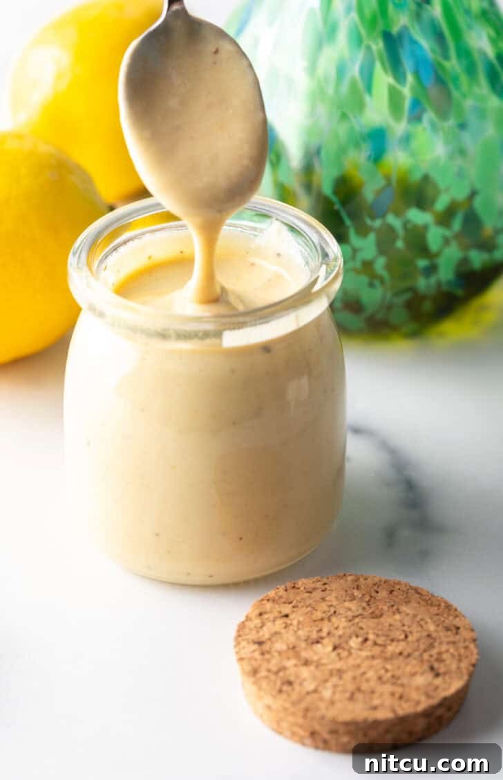 A spoon of creamy tahini dressing held above a jar of homemade tahini dressing, showcasing its rich texture.
