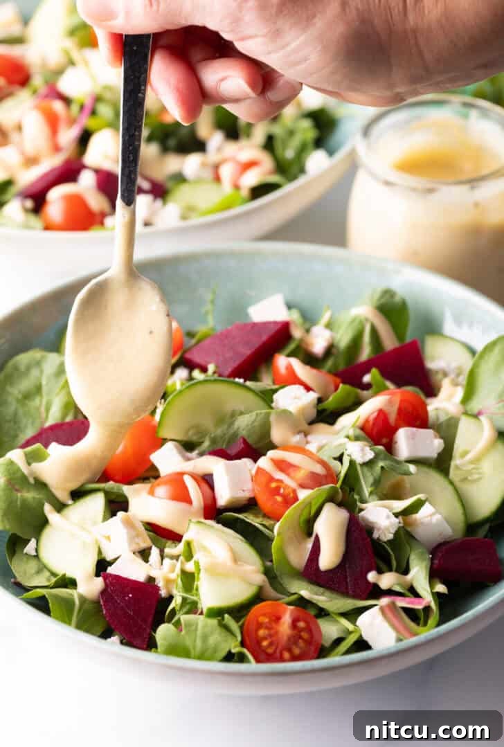 A hand with a spoon gently drizzling creamy tahini dressing onto a colorful Greek salad with cucumbers, mixed greens, tomatoes, and feta cheese cubes.