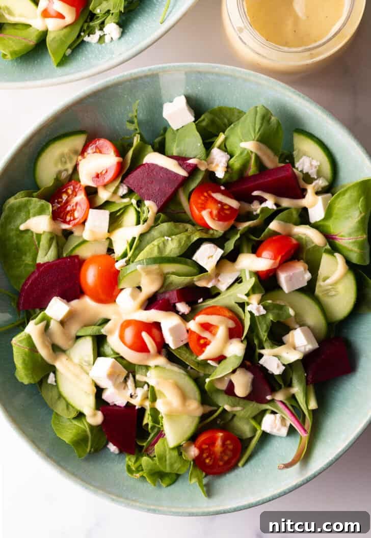 Top-down view of a colorful Greek salad with cucumbers, mixed greens, tomatoes, and feta cheese cubes, generously drizzled with pale yellow tahini dressing, showcasing its appetizing appeal.