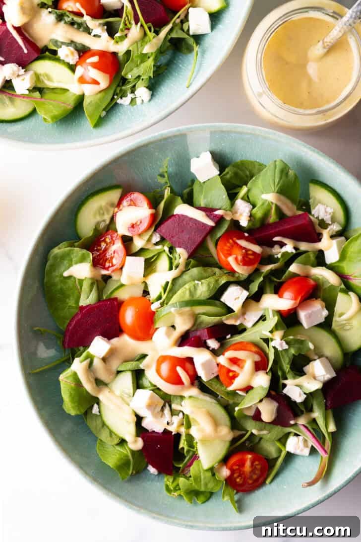 Top-down view of a colorful Greek salad with cucumbers, mixed greens, tomatoes, feta cheese cubes, lightly drizzled with pale yellow tahini dressing.