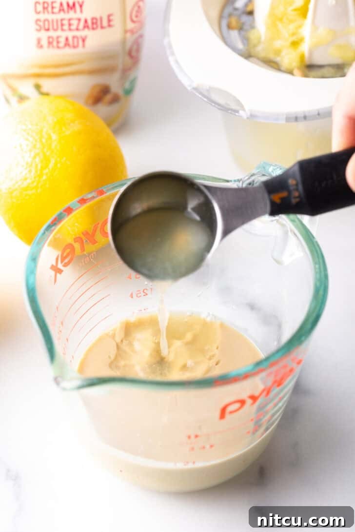 A hand adding fresh lemon juice from a small measuring spoon into a glass measuring jar containing tahini paste.
