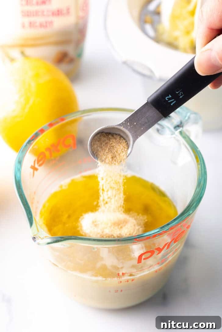 A hand adding spices from a small measuring spoon into a glass measuring jar with tahini paste and other wet ingredients.