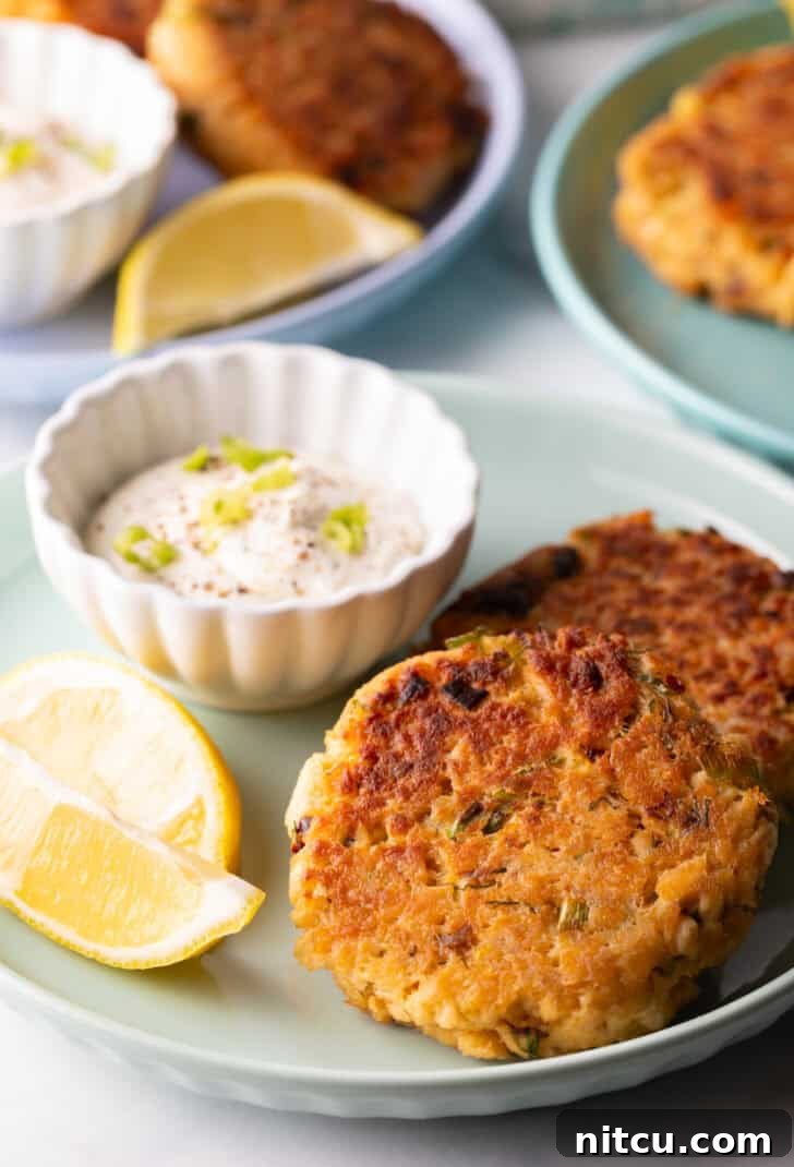 Effortless Golden Salmon Cakes 2 Two golden-brown salmon patties presented on a vibrant green plate, accompanied by a small white ramekin of creamy tartar sauce and two bright lemon wedges.