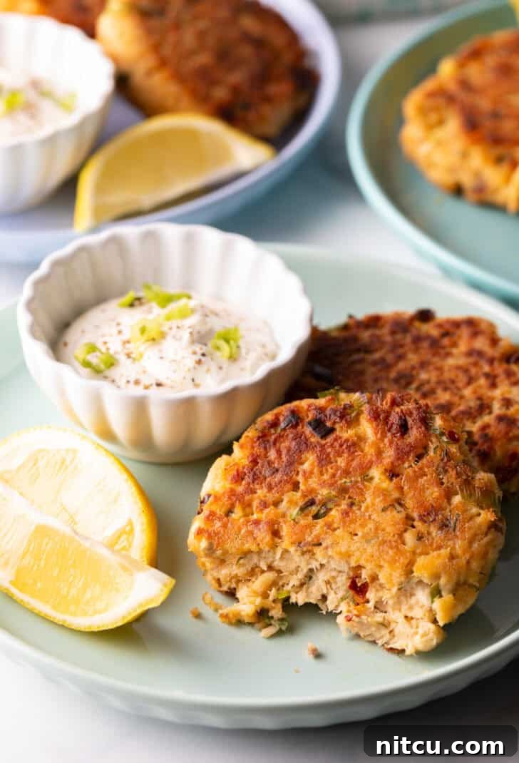 Effortless Golden Salmon Cakes 10 Two golden-brown salmon patties rest on a green plate, with a small white ramekin of tartar sauce and two lemon wedges. A noticeable bite has been taken from the patty closest to the camera, showcasing its flaky interior.