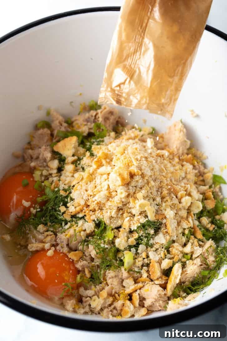 Effortless Golden Salmon Cakes 5 A large white mixing bowl containing flaked canned salmon, crushed crackers, vibrant fresh herbs, and cracked eggs, ready to be combined for salmon patties.