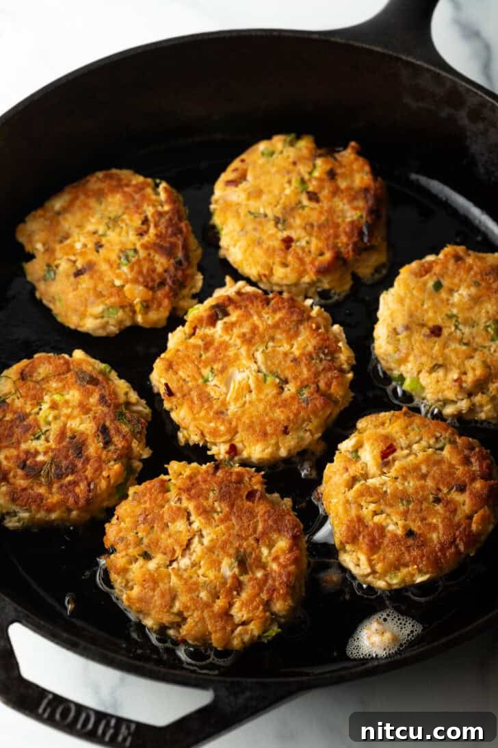 Effortless Golden Salmon Cakes 7 A top-down view captures seven salmon patties sizzling and browning beautifully in a cast iron skillet, illustrating the pan-frying process.