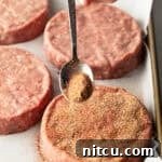 Sprinkling burger seasoning onto a raw hamburger patty with a metal spoon.
