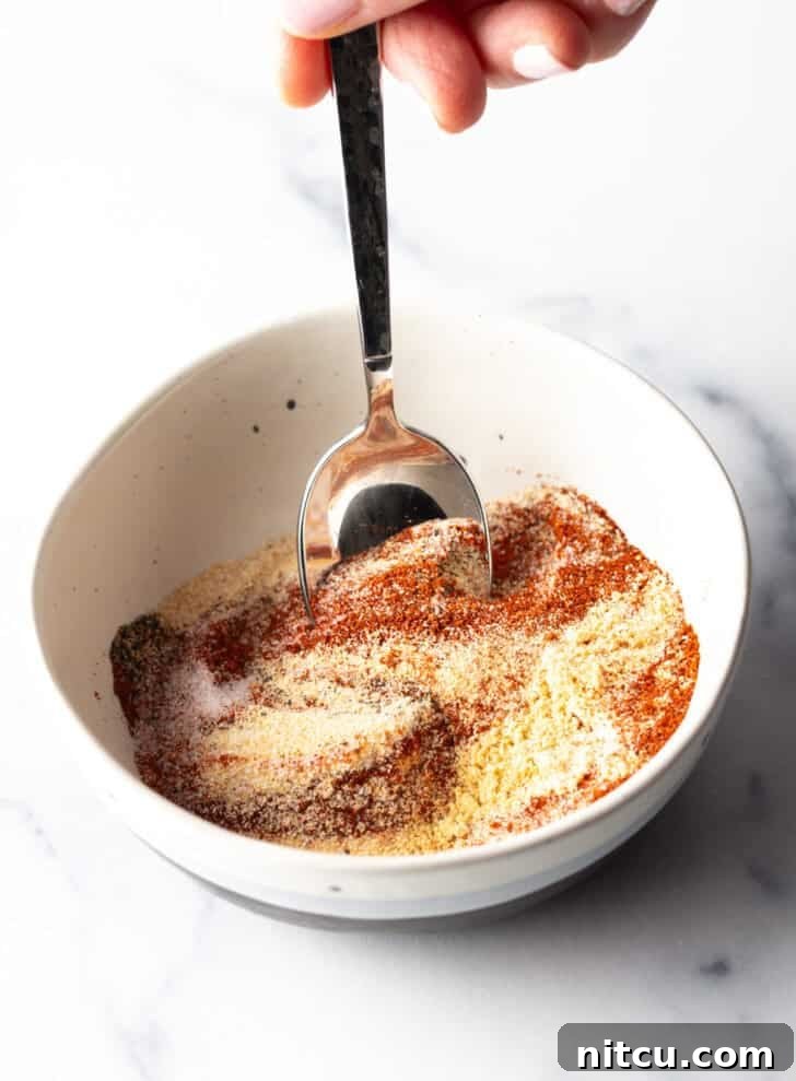The Ultimate Homemade Burger Spice Blend 6 A hand stirring together various spices in a white bowl with a spoon, showing the blending process of the burger seasoning.
