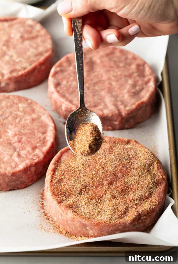 The Ultimate Homemade Burger Spice Blend 8 A metal spoon sprinkling a generous amount of homemade burger seasoning onto a raw hamburger patty.
