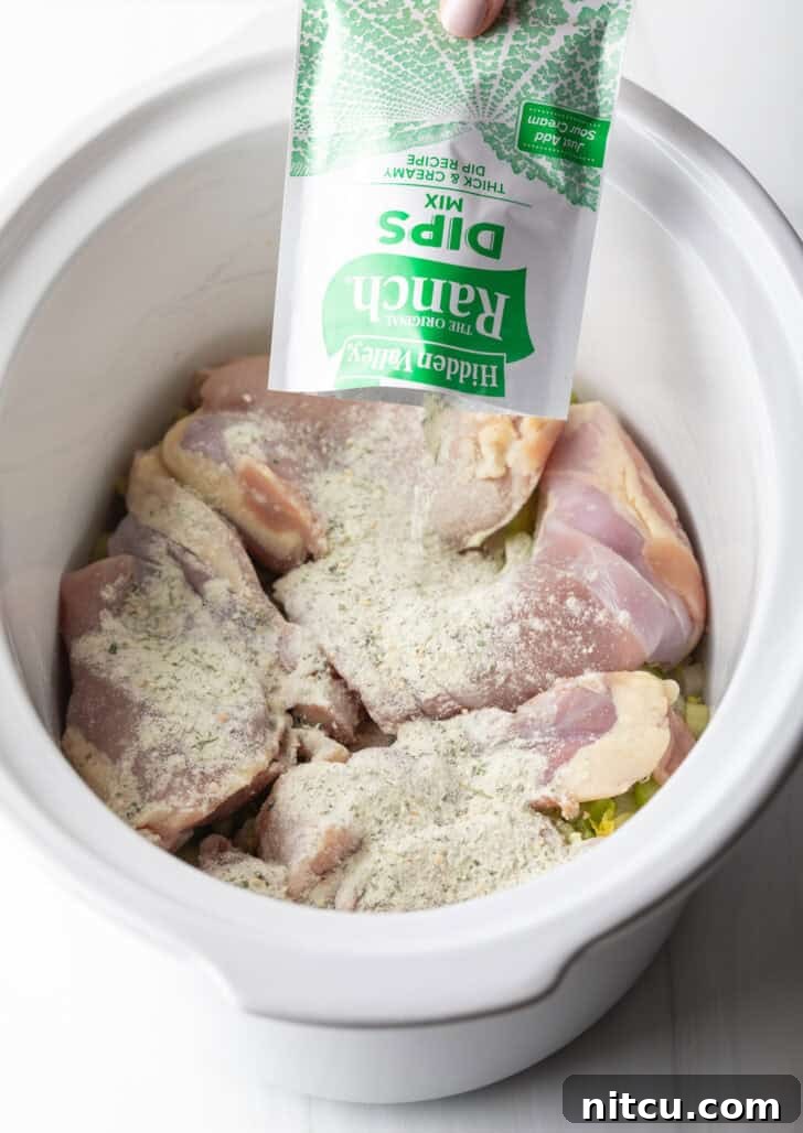 Sprinkling ranch dip powder over raw boneless, skinless chicken thighs in a white crockpot.