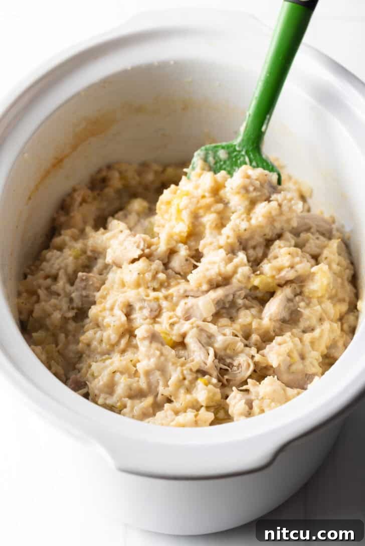 Cooked chicken and rice in a white crockpot, being stirred with a green spatula to show its creamy texture.