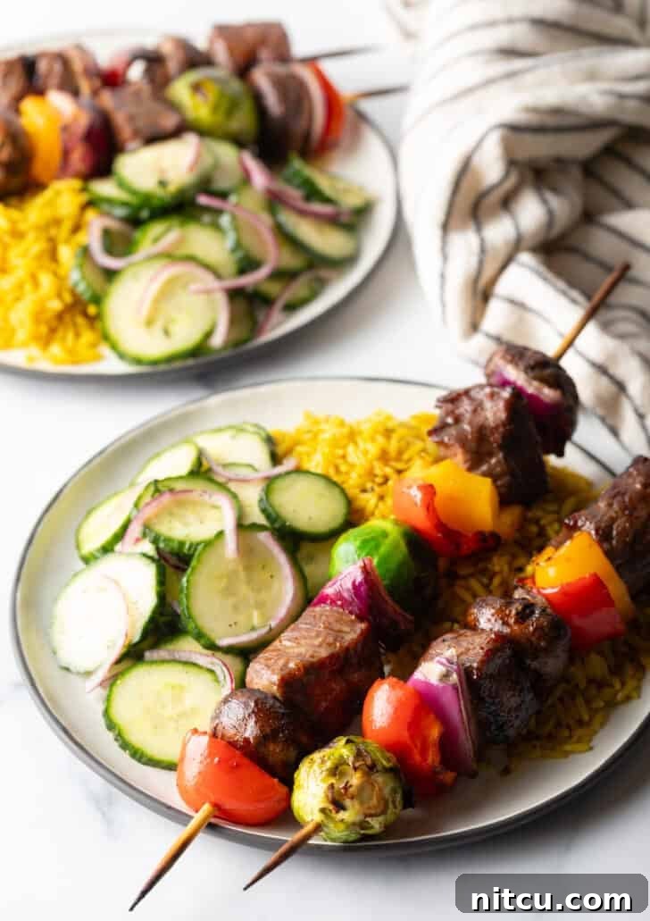 A beautifully presented white plate with two grilled beef and veggie kabobs, alongside a generous portion of yellow rice and a fresh cucumber salad.