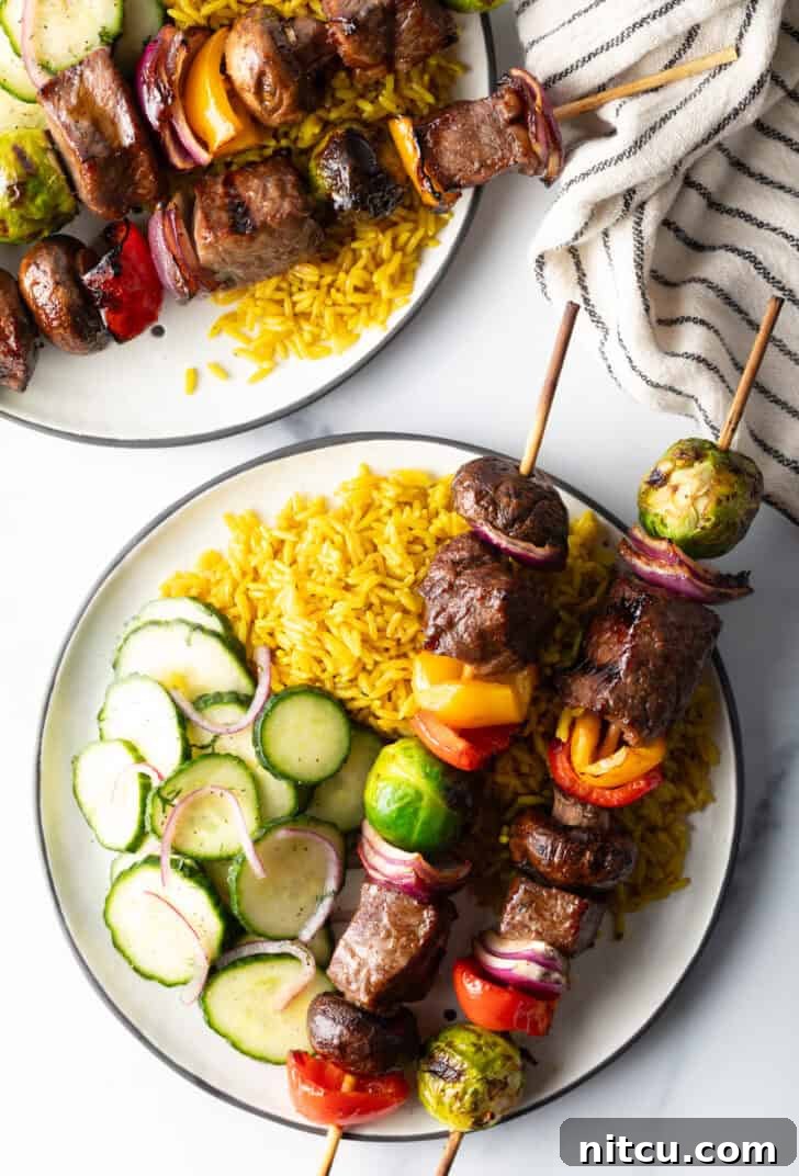 A elegant white plate featuring two succulent beef and veggie kabobs, perfectly complemented by a vibrant bed of yellow rice and a crisp cucumber salad.