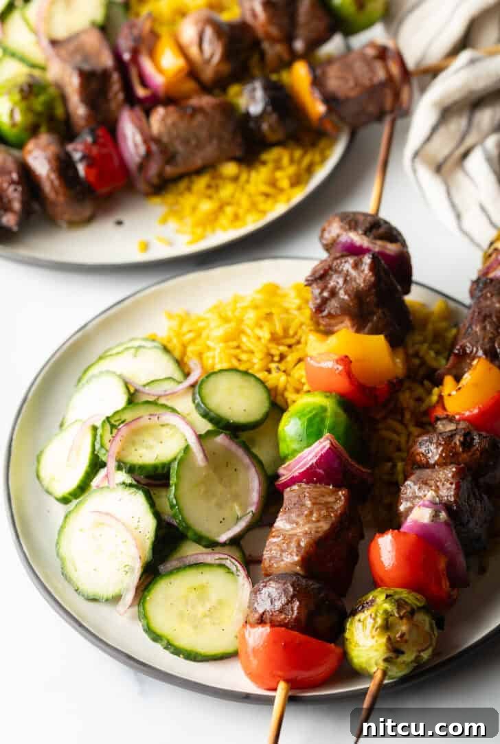 Close-up of a white plate holding two perfectly cooked beef and vegetable kabobs, with a blurred background of yellow rice and cucumber salad.