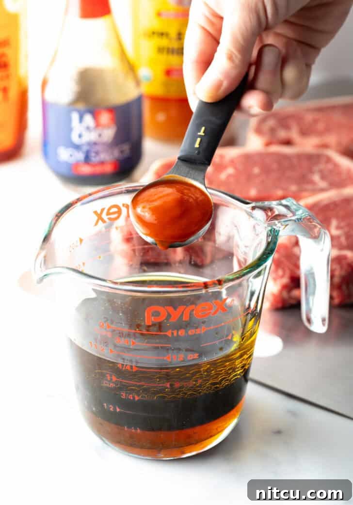 A hand adding a spoonful of ketchup to a glass measuring cup, preparing the marinade.