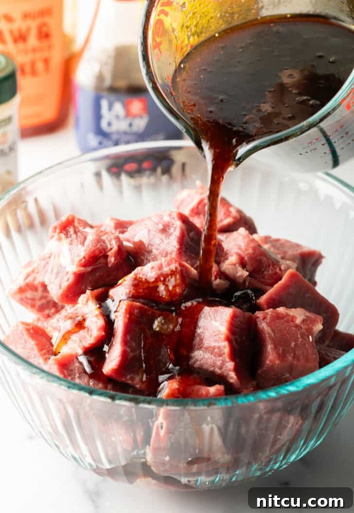 Marinade being poured from a glass measuring cup over perfectly cubed beef in a glass bowl.