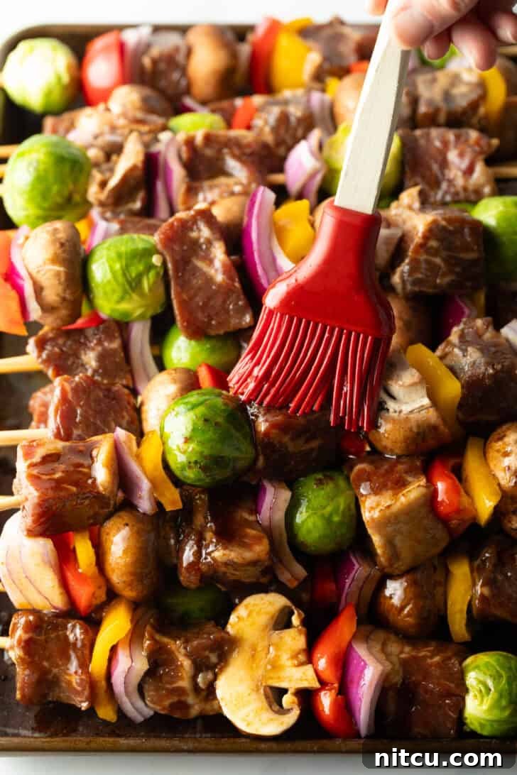 A red silicone brush basting a row of beef and vegetable kebabs on the grill, adding extra flavor.