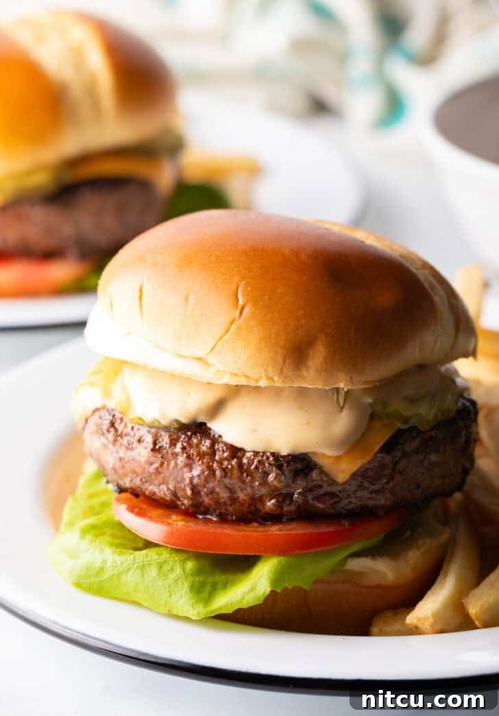 Signature Burger Dressing 4 A delectable cheeseburger featuring lettuce, tomato, melted cheese, and a generous layer of homemade burger sauce, served alongside crispy french fries on a white plate.