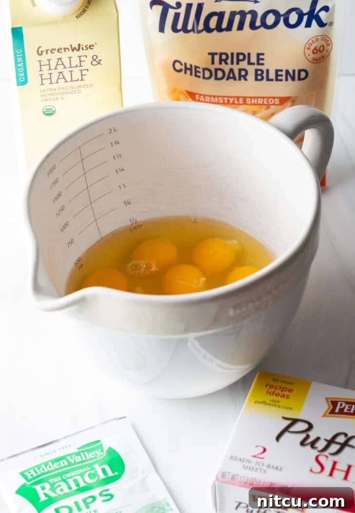 The Ultimate Effortless Breakfast Bake 5 Recipe ingredients neatly arranged: a large white mixing pitcher containing several eggs, a box of puff pastry, a packet of ranch dressing mix, a bag of shredded cheddar cheese, and a carton of half and half.