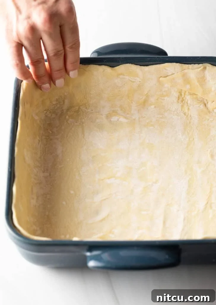 The Ultimate Effortless Breakfast Bake 7 A skilled hand gently placing the prepared puff pastry dough into the bottom and up the sides of a square baking dish, creating a perfect crust.