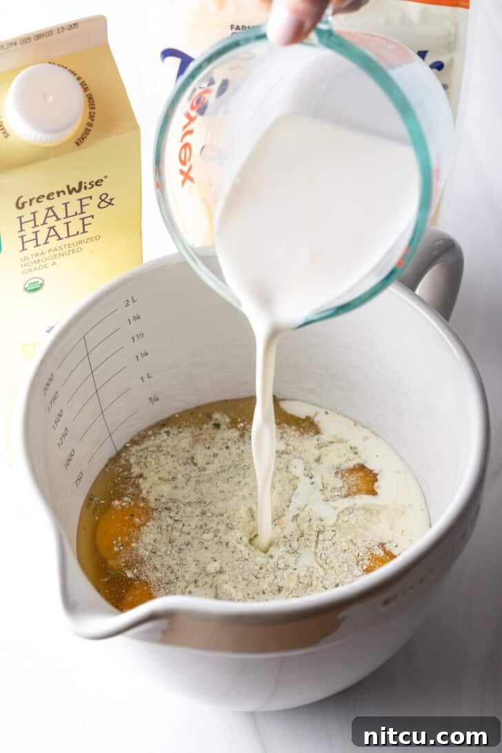 The Ultimate Effortless Breakfast Bake 8 Pouring creamy half and half from a clear glass measuring cup into a large white mixing pitcher already filled with cracked eggs, ready for whisking.