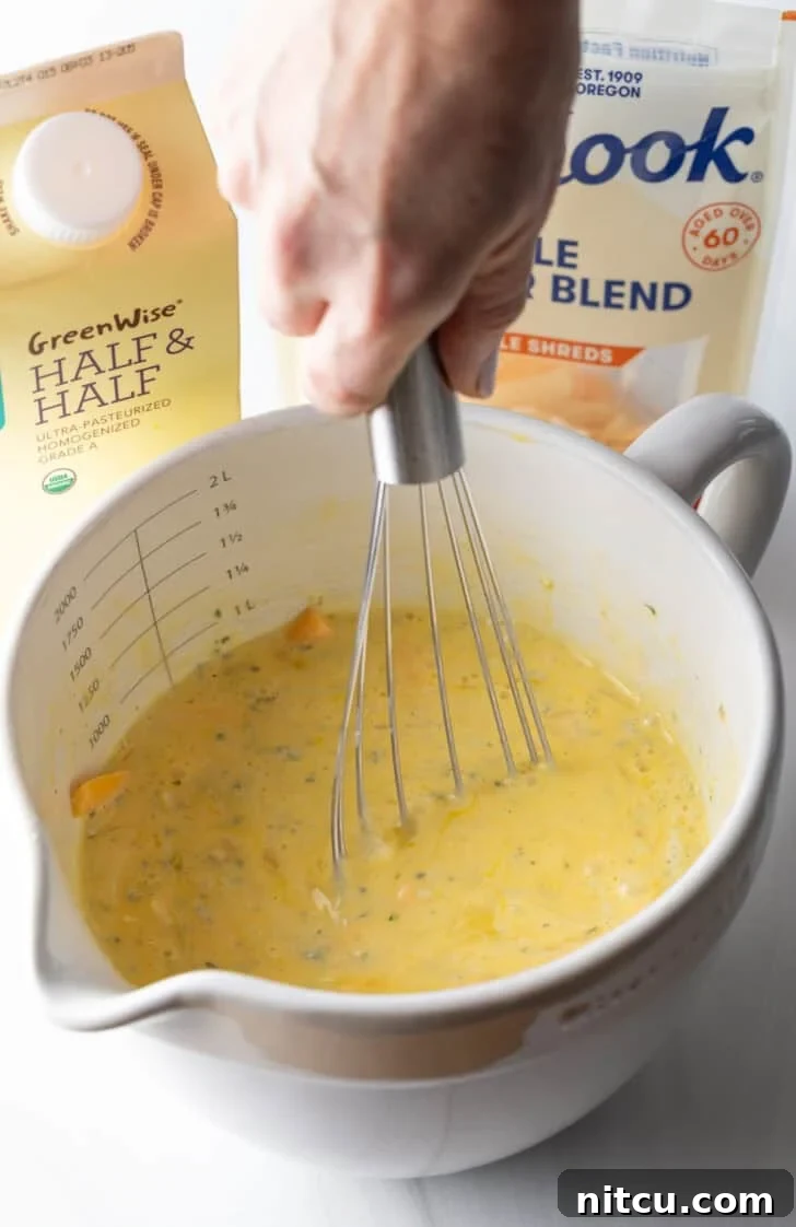 The Ultimate Effortless Breakfast Bake 9 A hand vigorously whisking eggs, half and half, and shredded cheese together in a large white mixing pitcher to create a smooth, well-combined egg mixture.