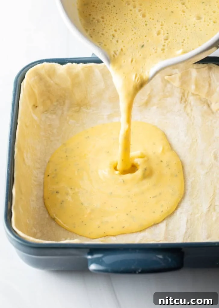 The Ultimate Effortless Breakfast Bake 10 Carefully pouring the perfectly blended egg mixture from a measuring cup into a prepared square baking dish, already lined with the delicate puff pastry crust.