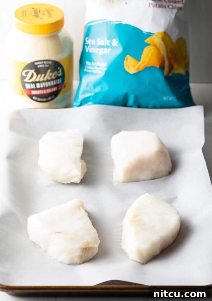 Four pieces of raw sea bass fillets arranged on white parchment paper, with a bag of salt and vinegar potato chips and a jar of mayonnaise in the soft background, showcasing the simple ingredients.