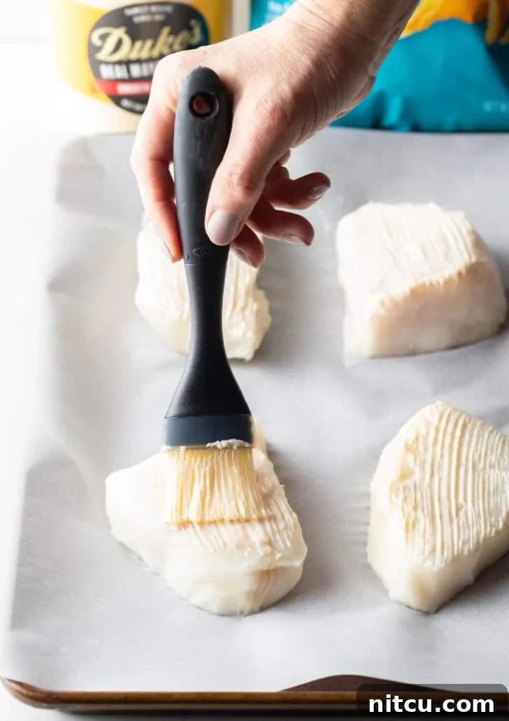 A black basting brush evenly coats four white grouper fillets with a thin layer of oil and seasonings on a baking sheet, preparing them for the next step.