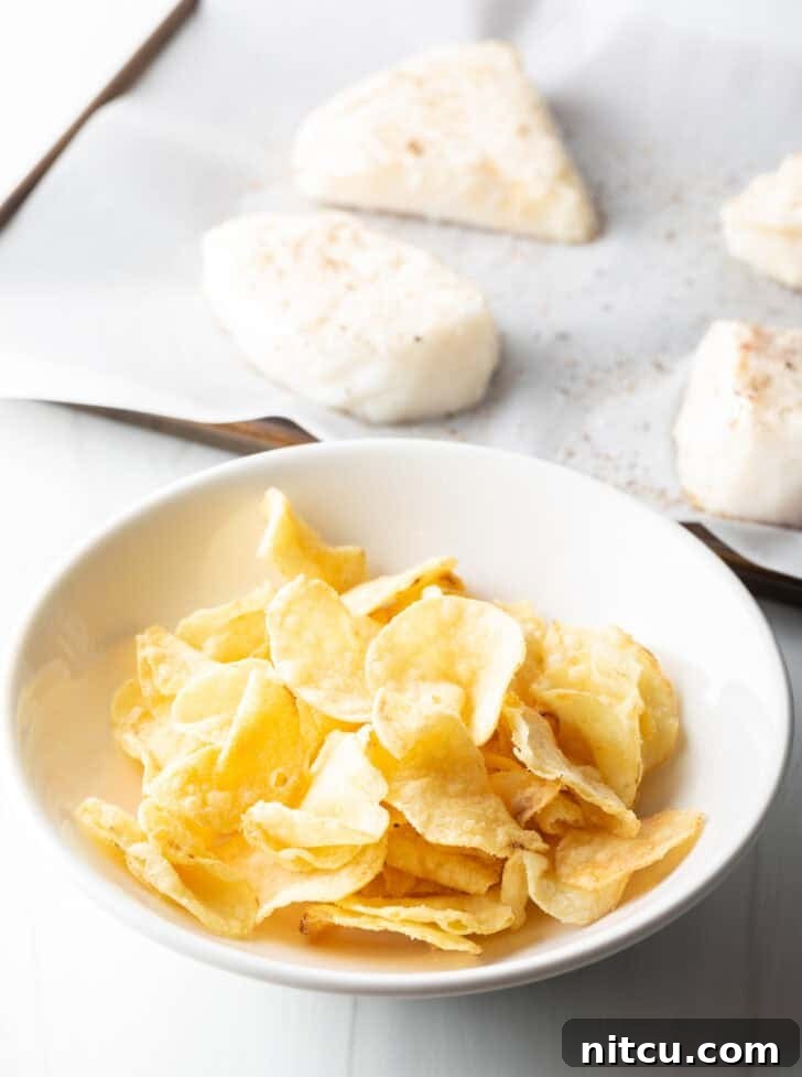 A white bowl filled with whole potato chips sits in the foreground, with four raw sea bass fillets waiting in the background, ready to be coated.