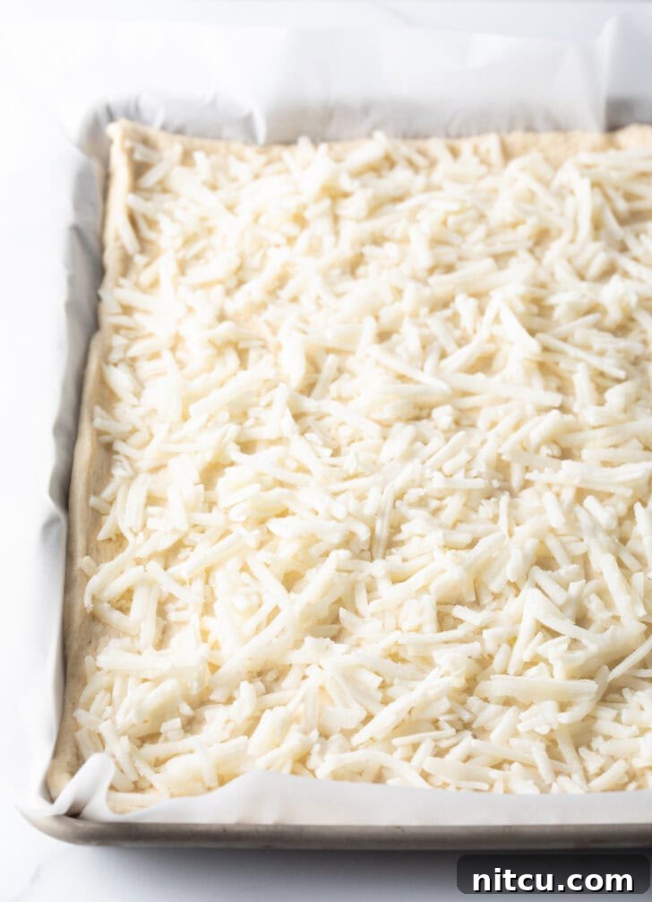 Daybreak Pizza 5 A close-up shot of an unbaked rectangular pizza crust, generously topped with a layer of shredded mozzarella cheese, ready for the oven.