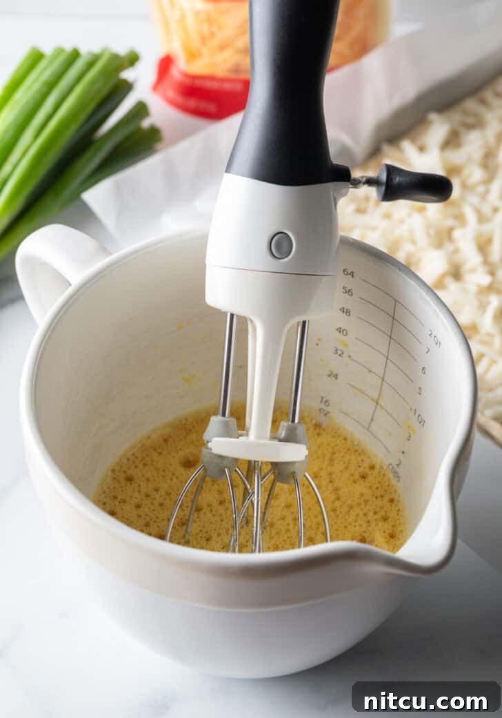 Daybreak Pizza 7 A white mixing pitcher containing a whisked egg mixture, with a hand holding a hand mixer, illustrating the preparation process for the fluffy eggs.