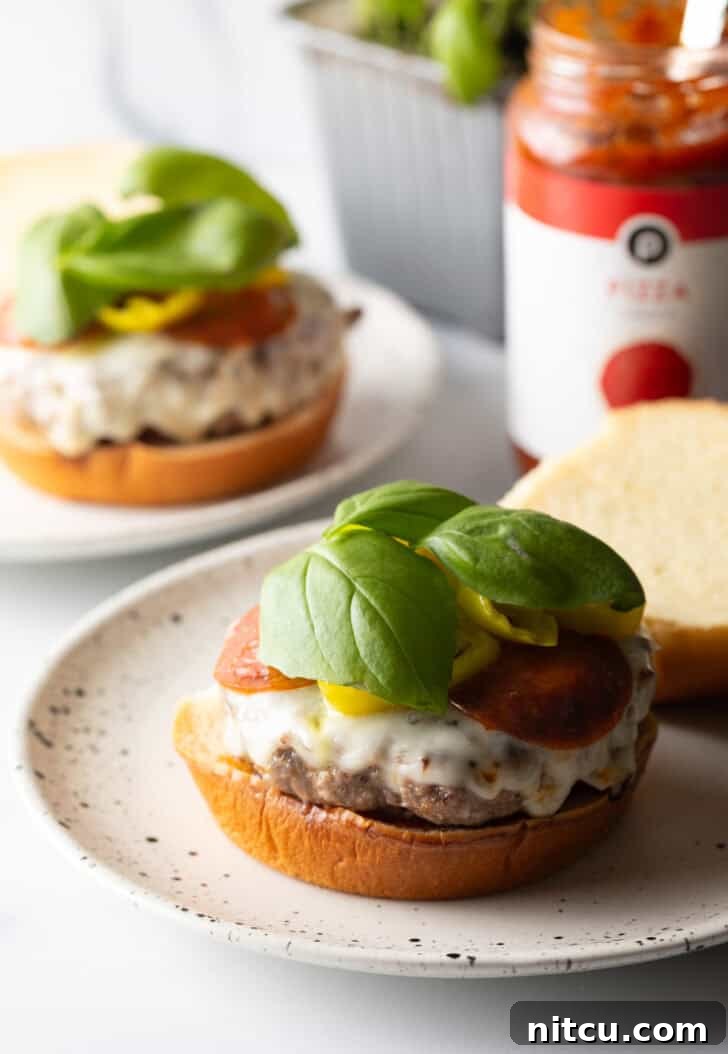 A beautifully composed open-faced burger on a white and black speckled plate, featuring the bottom bun, sauce, a stacked burger patty with melted mozzarella and pepperoni, artfully garnished with banana pepper rings and fresh baby basil leaves.