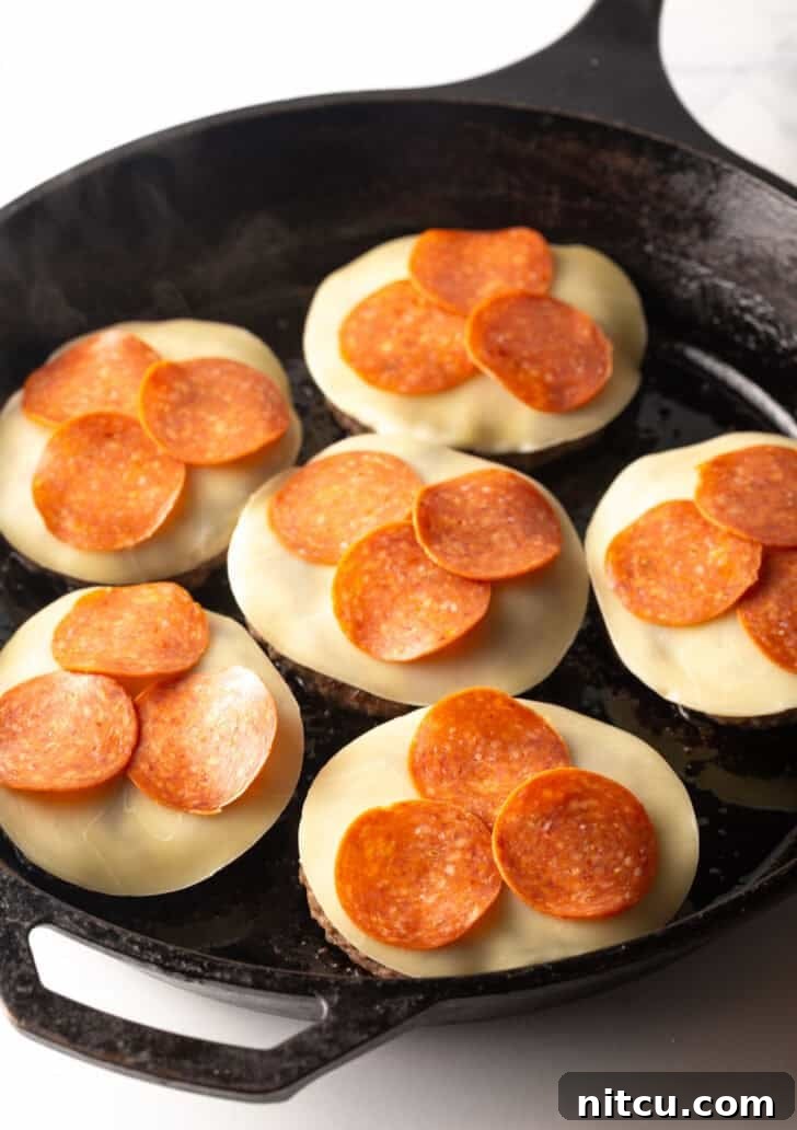 Six burger patties in a cast iron skillet, now topped with luscious mozzarella cheese slices and vibrant pepperoni slices, ready for melting.