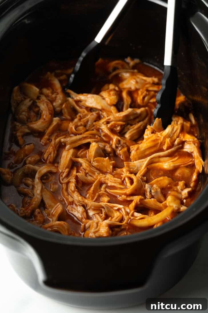 Tongs in a black crockpot with pulled BBQ chicken.