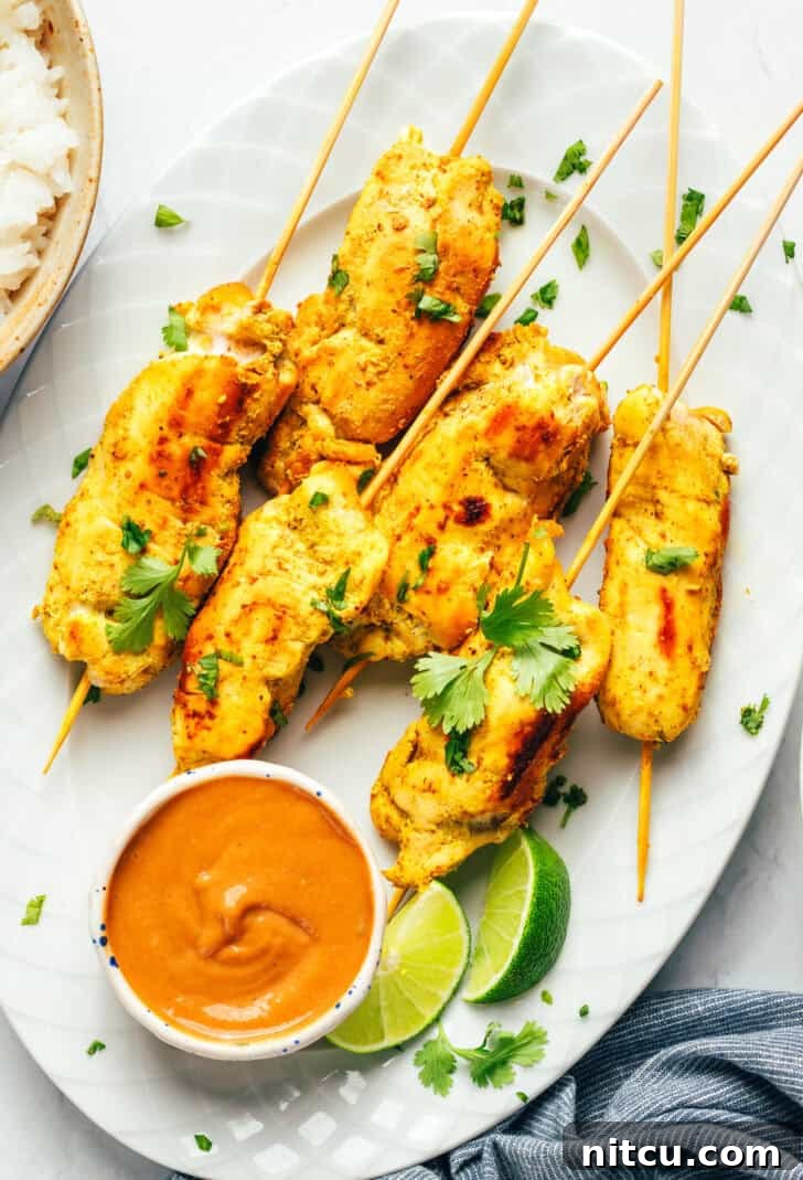 Sizzling Chicken Satay Skewers 2 Six pieces of golden-brown chicken satay elegantly arranged on a white oval plate, garnished with vibrant lime slices and accompanied by a small white ramekin filled with rich peanut sauce.