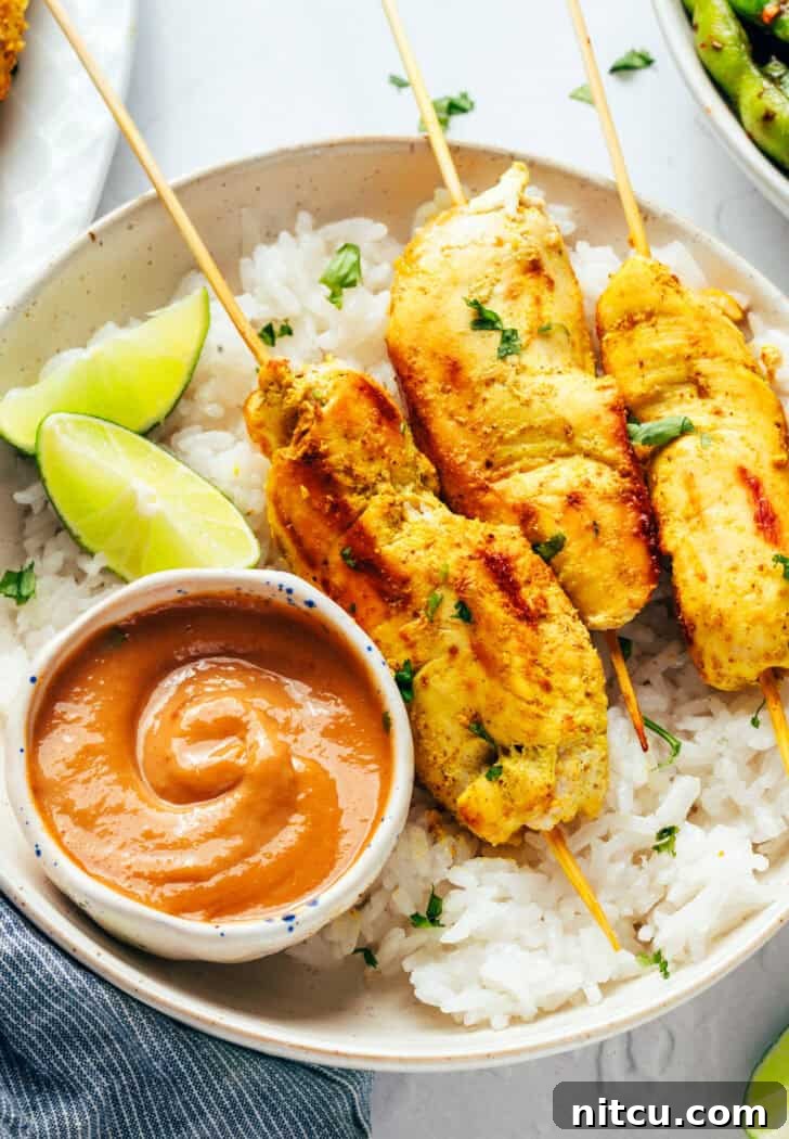 Sizzling Chicken Satay Skewers 13 Six pieces of chicken satay arranged on a white round plate with a side of fluffy white rice, vibrant lime slices, and a small white ramekin of rich peanut sauce.