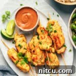 6 pieces of chicken satay on a white oval plate with slices of lime and a white ramekin of peanut sauce.